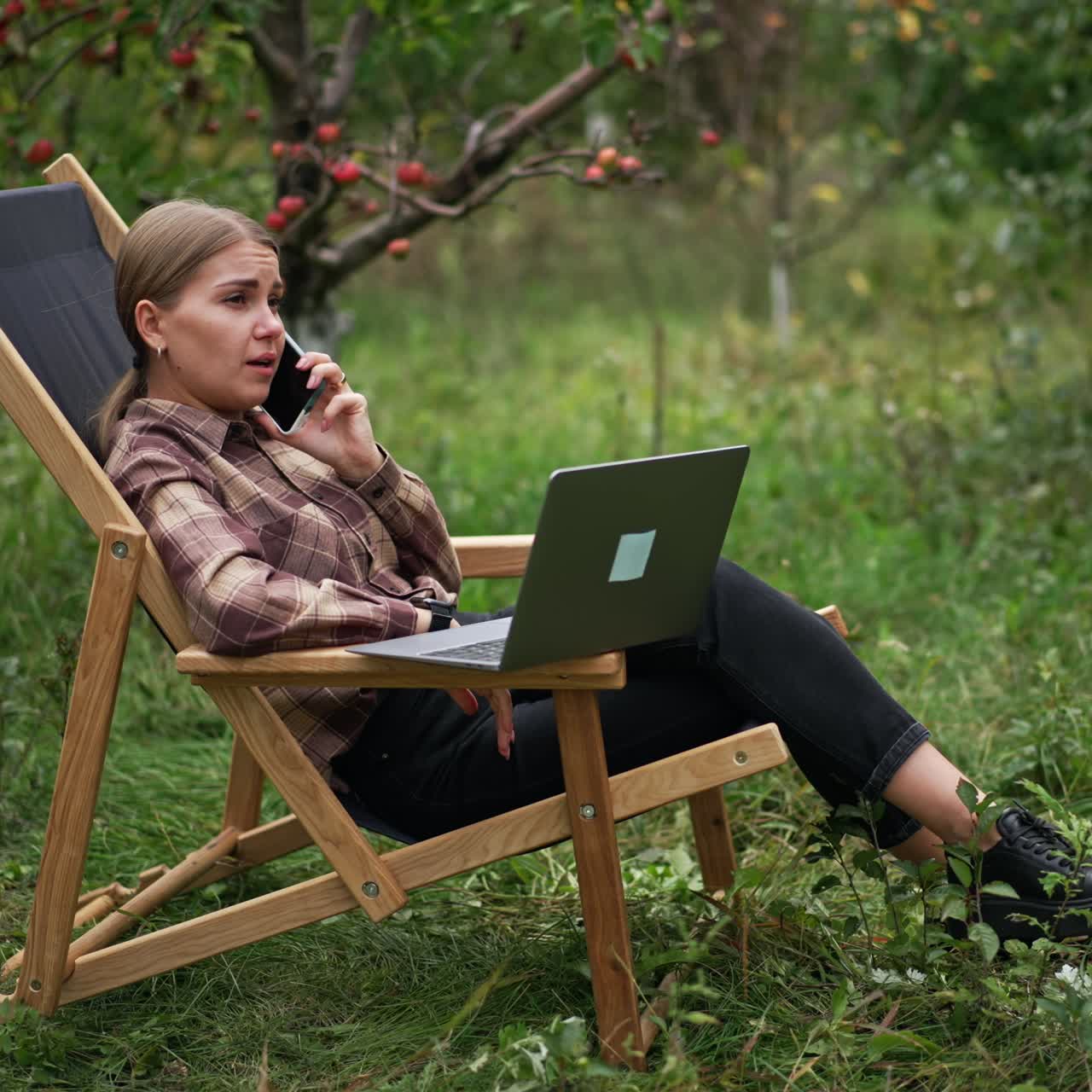 Phone conversation during the work remotely from office. Freelancer lady with laptop in front of her speaking cheerfully on the smartphone. Garden backdrop