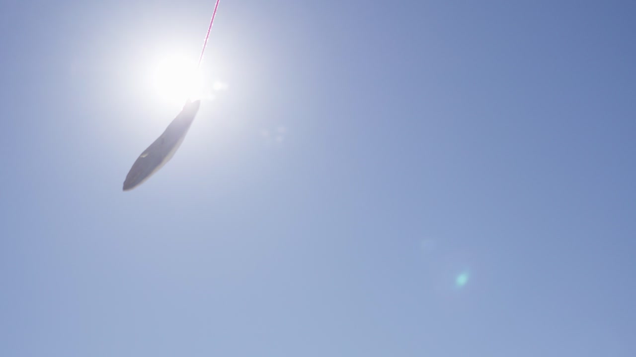A single sand dollar sways on a red string in the breeze, sunlight glowing through its delicate shell against a wide clear sky, capturing a serene coastal vibe.