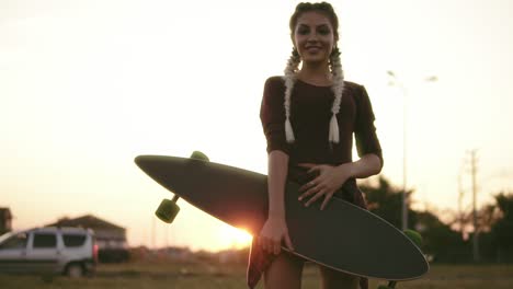 Junge-Attraktive-Frau-In-Kariertem-Hemd,-Shorts-Und-Tanktop,-Die-Longboard-Hält-Und-Während-Des-Sonnenuntergangs-Im-Sommer-In-Die-Kamera-Schaut