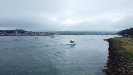 Motorboat-rising-aerial-view-entering-Welsh-seaside-town-harbour-cruising-along-River-Conwy