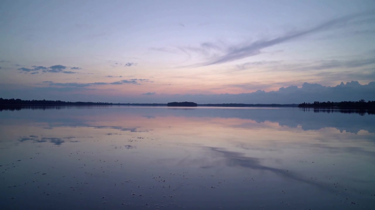 Wind stirring clouds nudging calm lake water at dusk, showing island, horizon, trees, dark specks