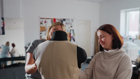 clothing designers exchange high five while adjusting beige waistcoat on black mannequin, mirror in background reflecting celebratory gesture, fabric rolls and mood board filling bright sewing studio