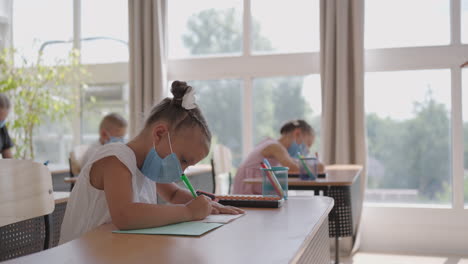 student in protective mask studying at school during corona virus pandemic. a masked teacher explains a new lesson topic to masked students. children write with pens in the classroom