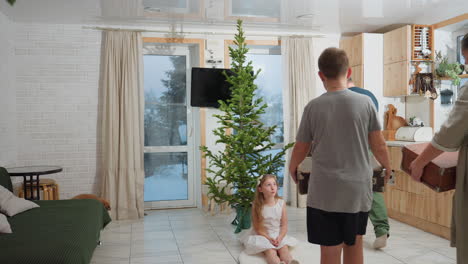 Little girl in white gown sitting on rug in front of decorated Christmas tree, as family member approaches with box for holiday surprise or gift exchange