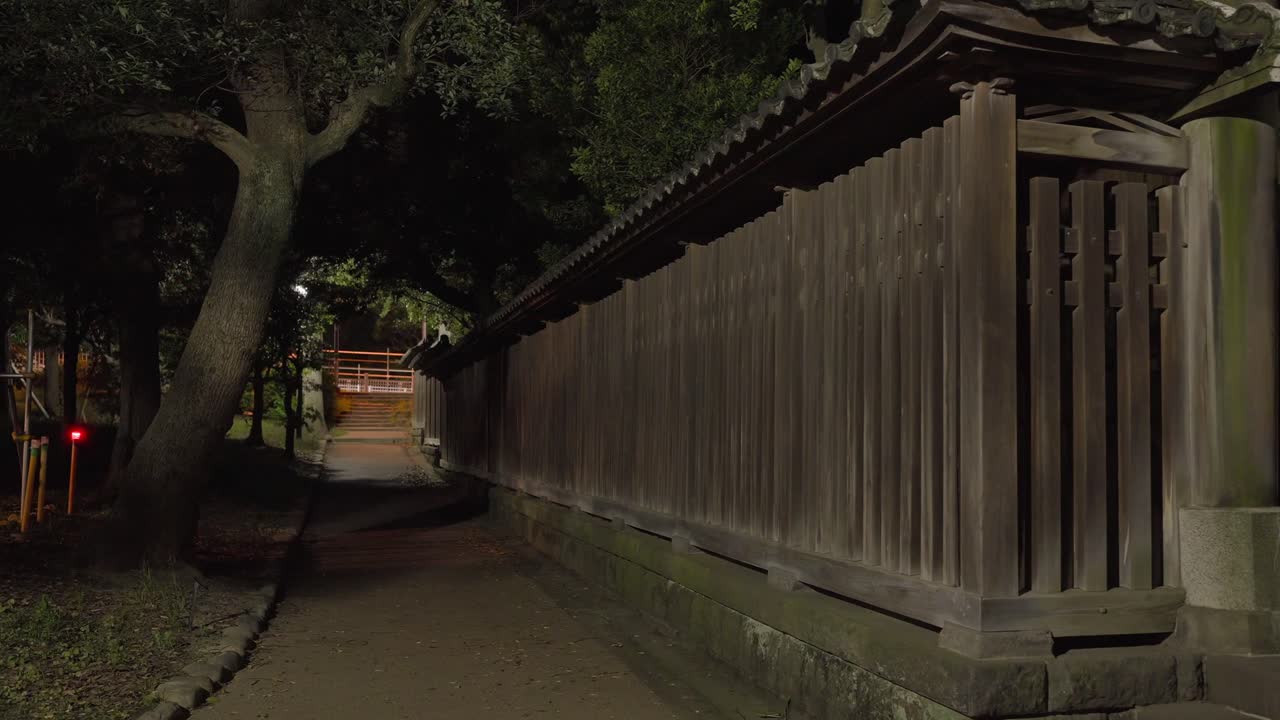 A serene night shot of a traditional Japanese wooden fence and a quiet path, with a large tree in the background