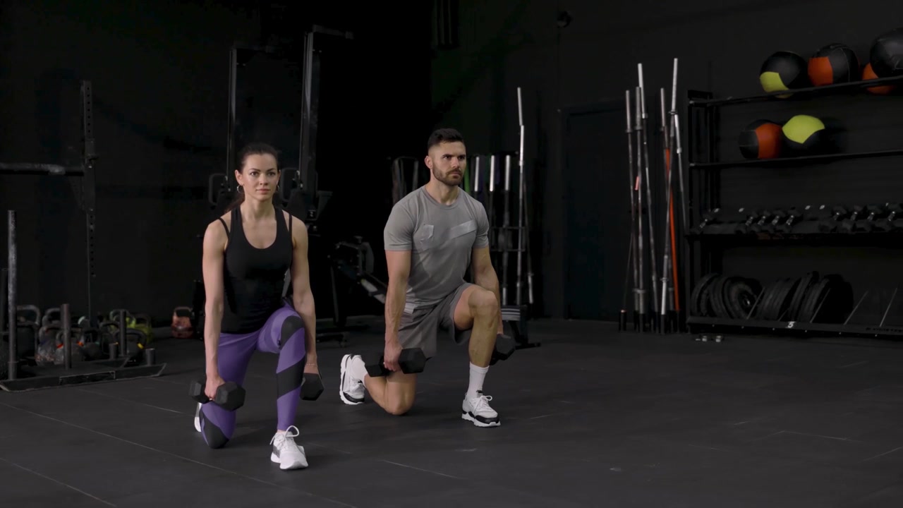 Hombre y mujer realizando zancadas con mancuernas juntos en un gimnasio