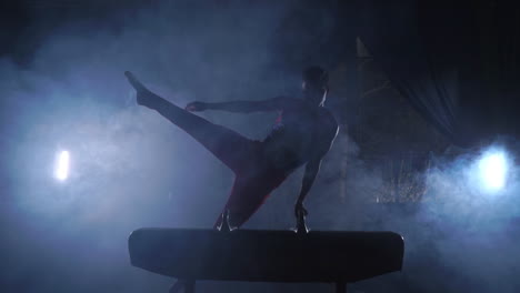 male gymnast performs exercises from the olympic program on pommel horse performing spins and rolls in slow motion getting ready for the olympics by training hard in the dark room around the smoke and the counter light