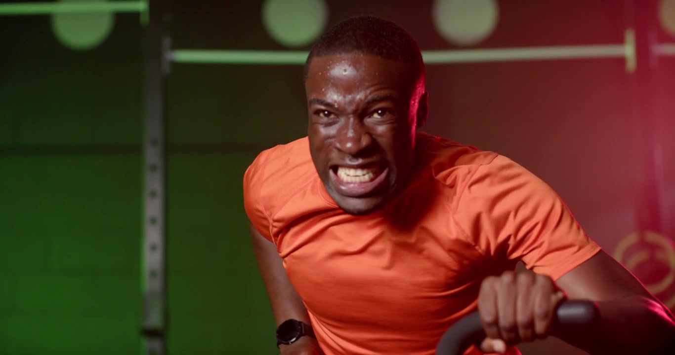 Adult African man gripping cardio bike handles, pulling for interval training in gym in orange tee