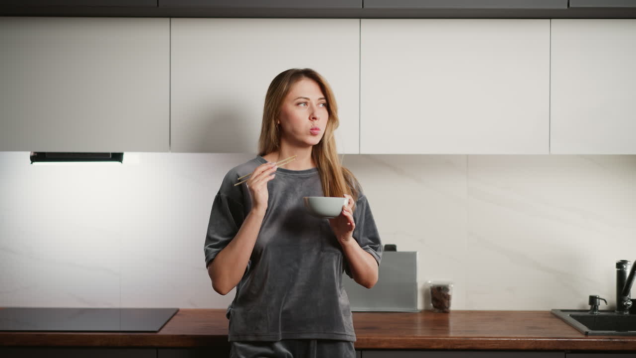 Young lady standing in kitchen eating spaghetti with chopsticks, capturing casual indoor dining moment with relaxed vibe, warm lighting, and modern home environment