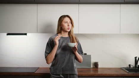 young lady standing in kitchen eating spaghetti with chopsticks, capturing casual indoor dining moment with relaxed vibe, warm lighting, and modern home environment