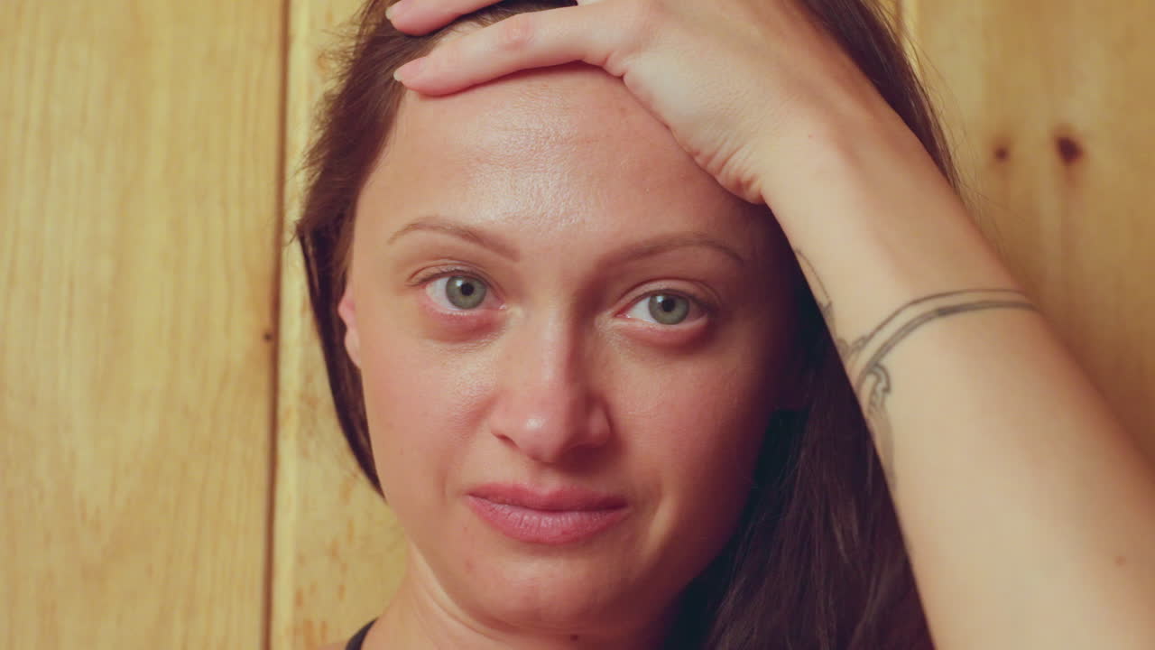 Close up of serene woman resting hand on forehead while smiling gently, eyes closed in peaceful sauna setting, soft lighting highlighting facial expression and subtle tattoo detail on relaxed arm