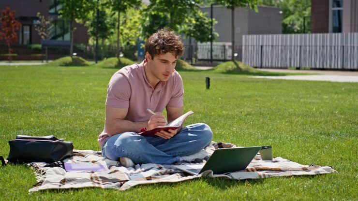 Studente online seduto sull'erba in un parco mentre fa una videochiamata dal portatile. Uomo rilassato