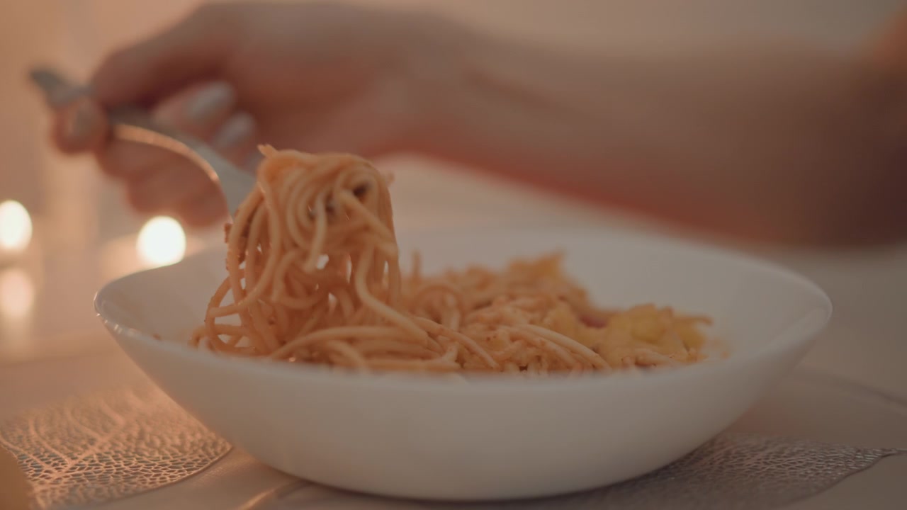 Un primo piano caldo mostra una forchetta che mescola spaghetti cotti in una ciotola bianca su un tavolo illuminato da candele, con la mano visibile e un delicato movimento dei noodles che crea un'atmosfera da pranzo accogliente, bokeh morbido, atmosfera intima, e.