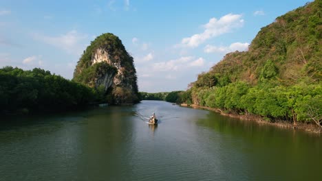 aerial drone flying tracking a thai longtail boat down river with large green limestone rocks and mangrove forests in the background on a sunset afternoon in krabi town thailand