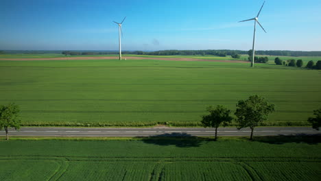 Toma-Aérea-De-Camiones-Eléctricos-Conduciendo-Por-Carreteras-Rurales-Y-Girando-Turbinas-Eólicas-En-El-Fondo---Hermoso-Día-Soleado-Con-Cielo-Azul-En-El-Campo