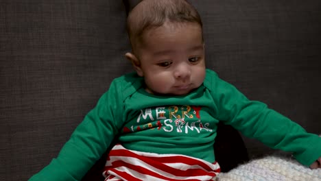 adorable bangladeshi baby wearing festive green merry christmas outfit sat on sofa looking side to side