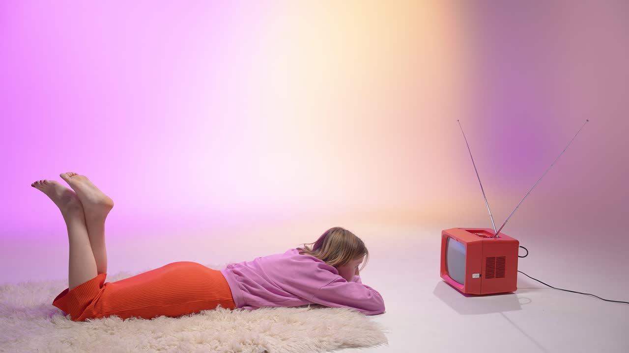 A young woman lies on a fluffy rug in front of a red vintage TV, bathed in soft pastel lighting. The scene exudes calm nostalgia, blending retro aesthetics with serene studio elegance
