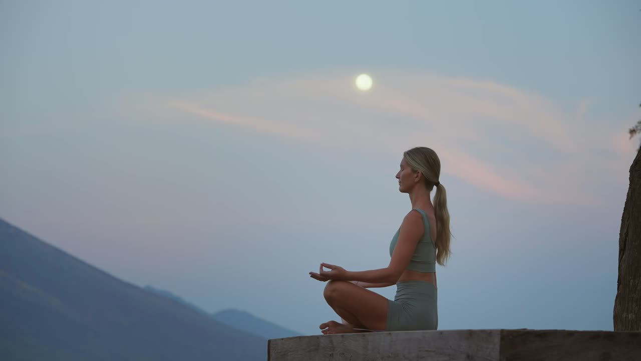 mujer meditando en una postura de yoga sentada en una plataforma de madera con luna llena