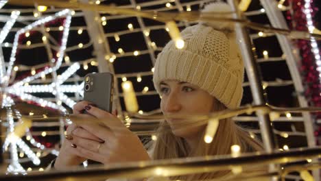 mujer con abrigo fotografiando coloridas decoraciones navideñas con un teléfono por la noche