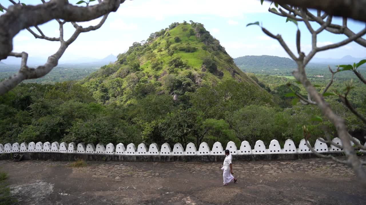 Buddhist man in white traditional clothes walks downhill with the high green hills of nature of Sri Lanka in Dambulla in the background. Wide shot through the branches