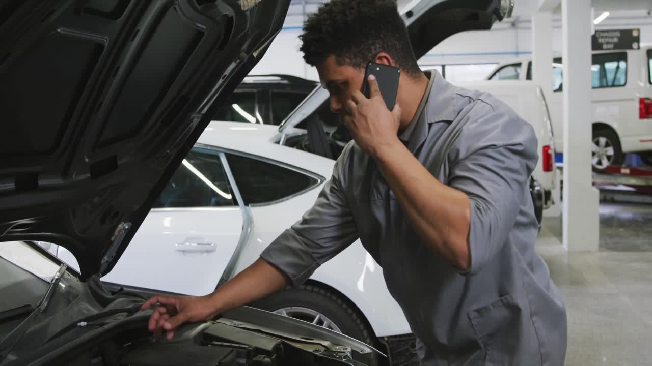 mecánico de automóviles afroamericano mirando un motor de automóvil abierto y hablando en un teléfono inteligente