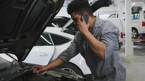 mecánico de automóviles afroamericano mirando un motor de automóvil abierto y hablando en un teléfono inteligente