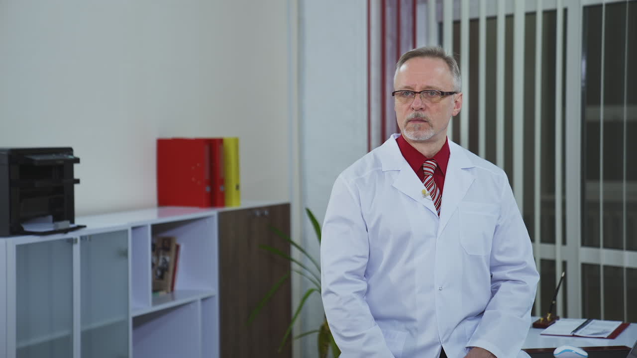 Médico sênior sentado à mesa do consultório e olhando para o telefone. Médico experiente e pensativo consulta paciente por videochamada.