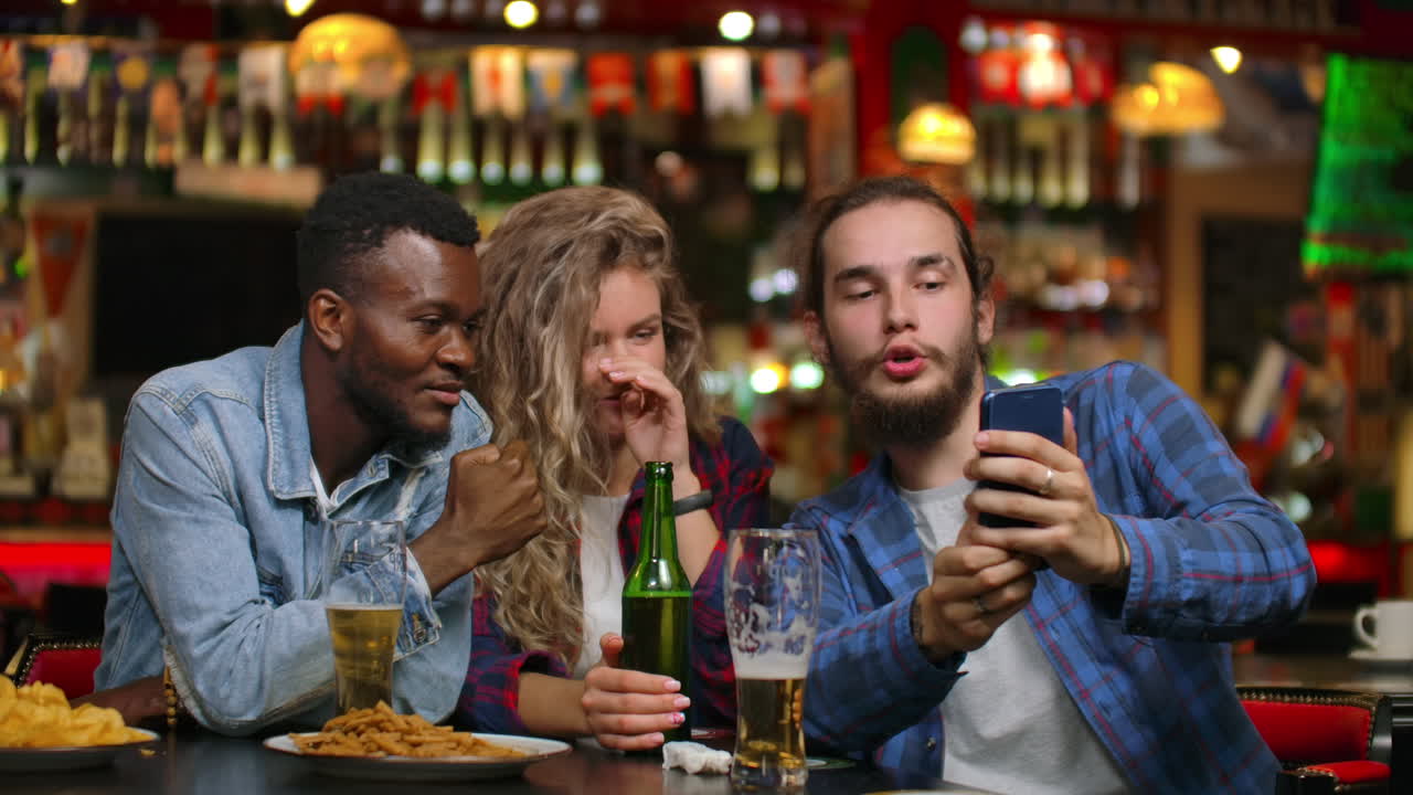 Two men and a woman look at the phone screen while sitting in a bar and prepare to take a shared selfie. Cheerful youth company at the bar.