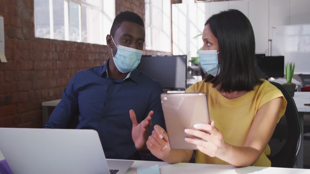 Diverse male and female business colleagues wearing face masks sitting at desk using tablet