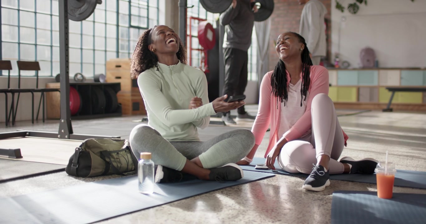 African-American female friends left woman scrolling phone showing content in gear laughing on mats