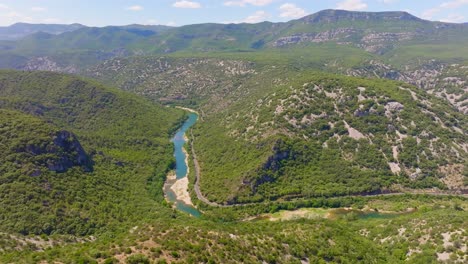 aerial drone shot over hills with a river running through the middle, mountains in the background, greenery and trees everywhere, blue sky and a few clouds