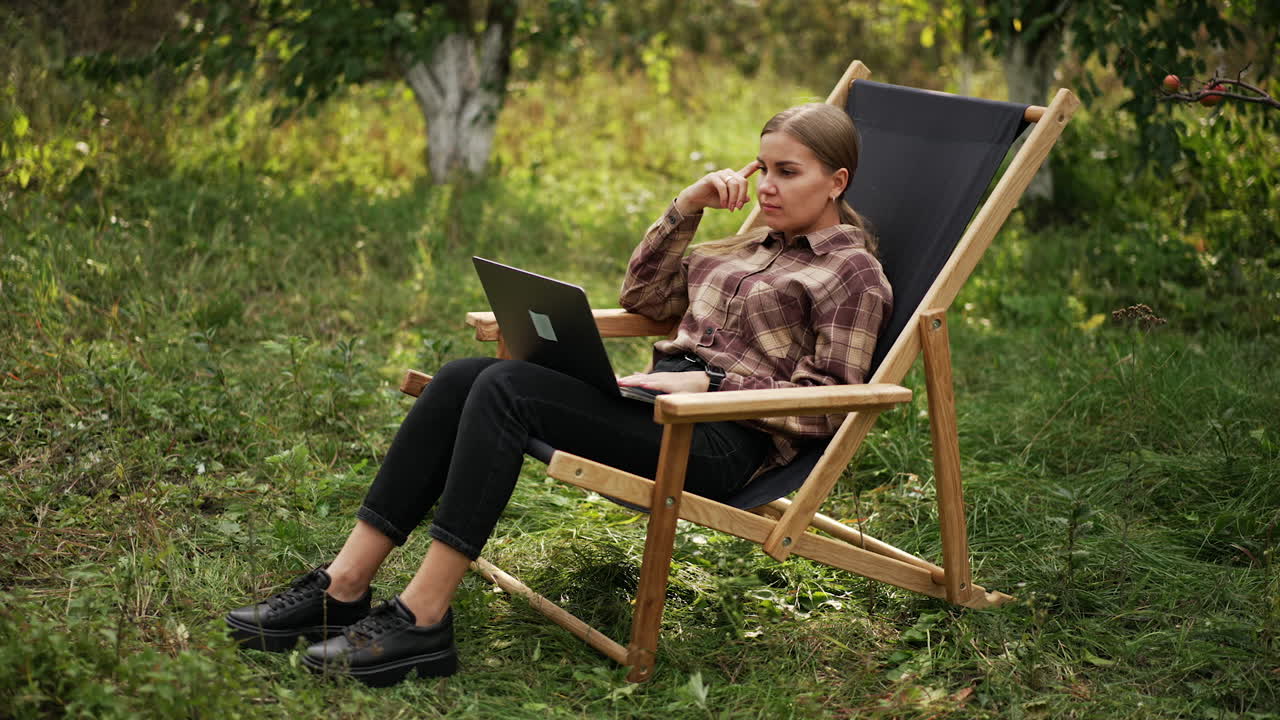 Freelancing young lady working on laptop in the garden. Lady stops for a moment to think over something and then continues work happily.