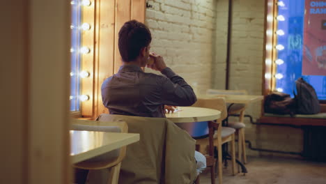 student in formal shirt sits alone inside cozy cafe near bright window lights, holding coffee cup while facing forward, with bottle on table, jacket draped on chair, and bags resting on window bench nearby