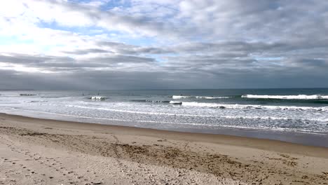 Ein-Wunderschöner-Sauberer-Sandstrand-Mit-Wellen-In-Zeitlupe-Und-Dramatischen-Wolken,-Durch-Die-Die-Sonne-Scheint