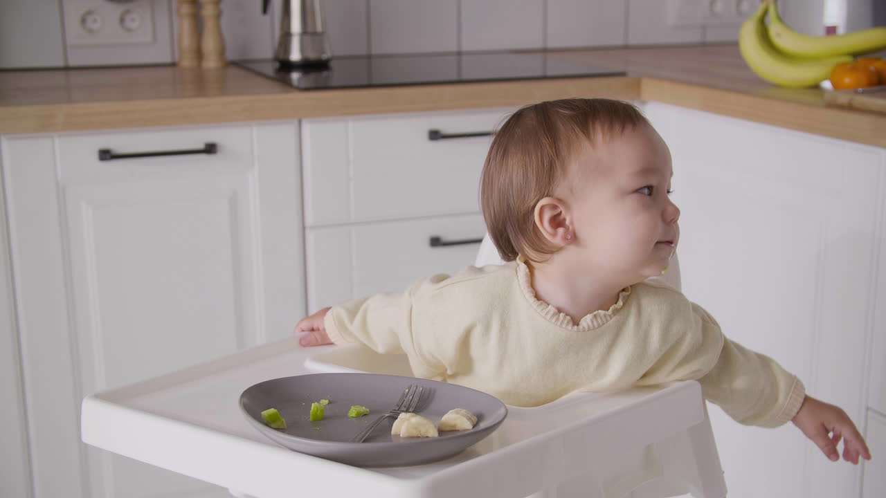 niña feliz sentada en una silla alta en la cocina, riendo y señalando algo durante la comida 1