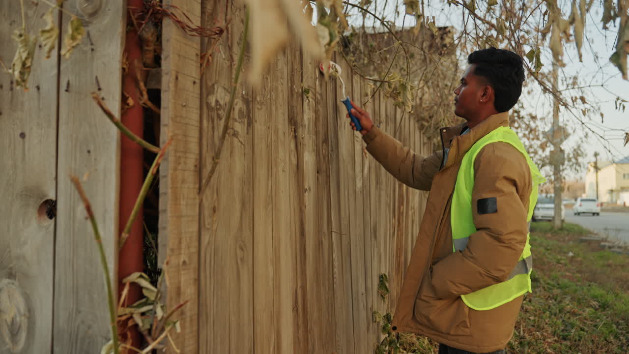 Operaio di colore che dipinge una staccionata di legno invecchiata, indossando un giubbotto ad alta visibilità, usando un rullo per riparare le assi danneggiate. Foglie autunnali, una strada di quartiere tranquilla, routine di manutenzione focalizzata.