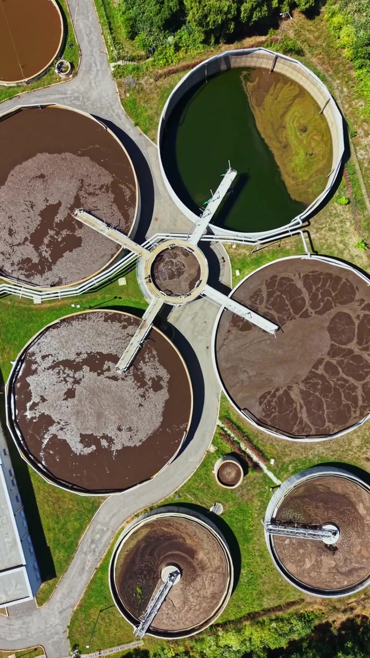 Water treatment facility processes wastewater in an aerial view showing multiple circular tanks and surrounding greenery