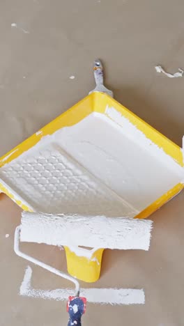 Vertical close-up showing two white paint rollers and a yellow tray filled with white paint, highlighting a renovation process involving wall or ceiling painting preparation