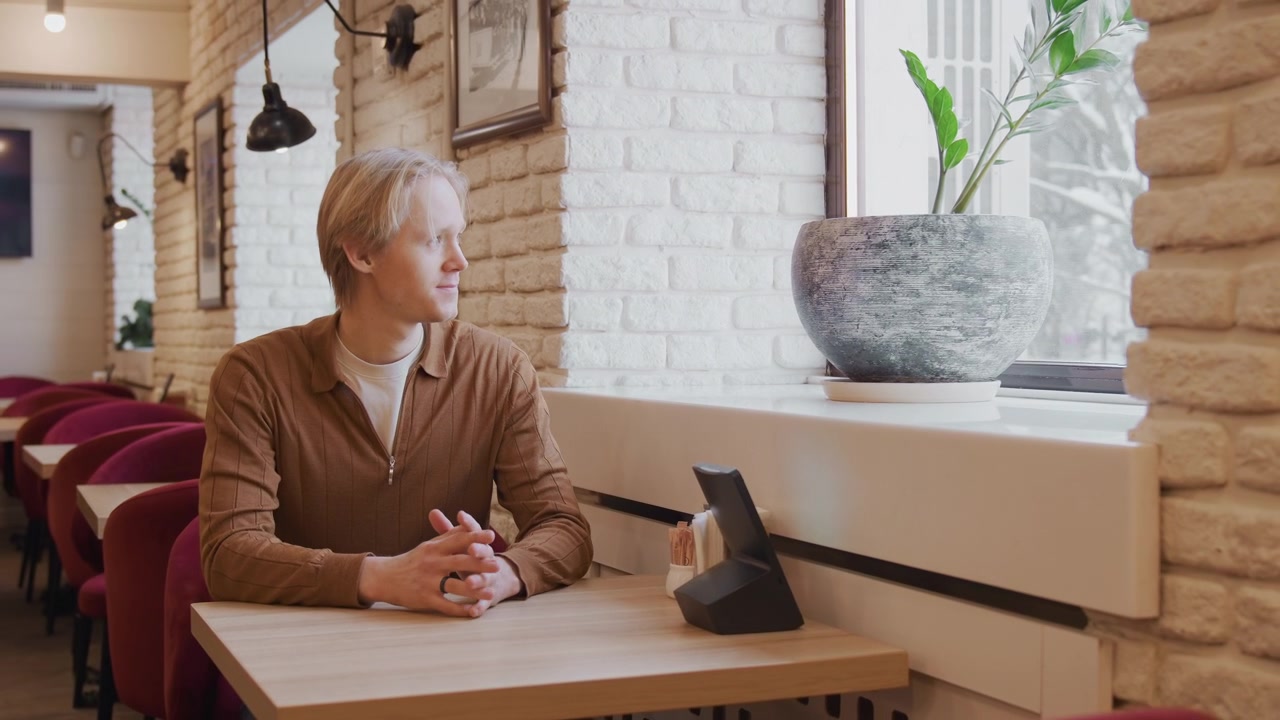 Man in brown sweater moves to sit at empty table with red chairs and white brick walls. Man adjusts chair places his hands on wooden tabletop near condiment holders like salt shakers