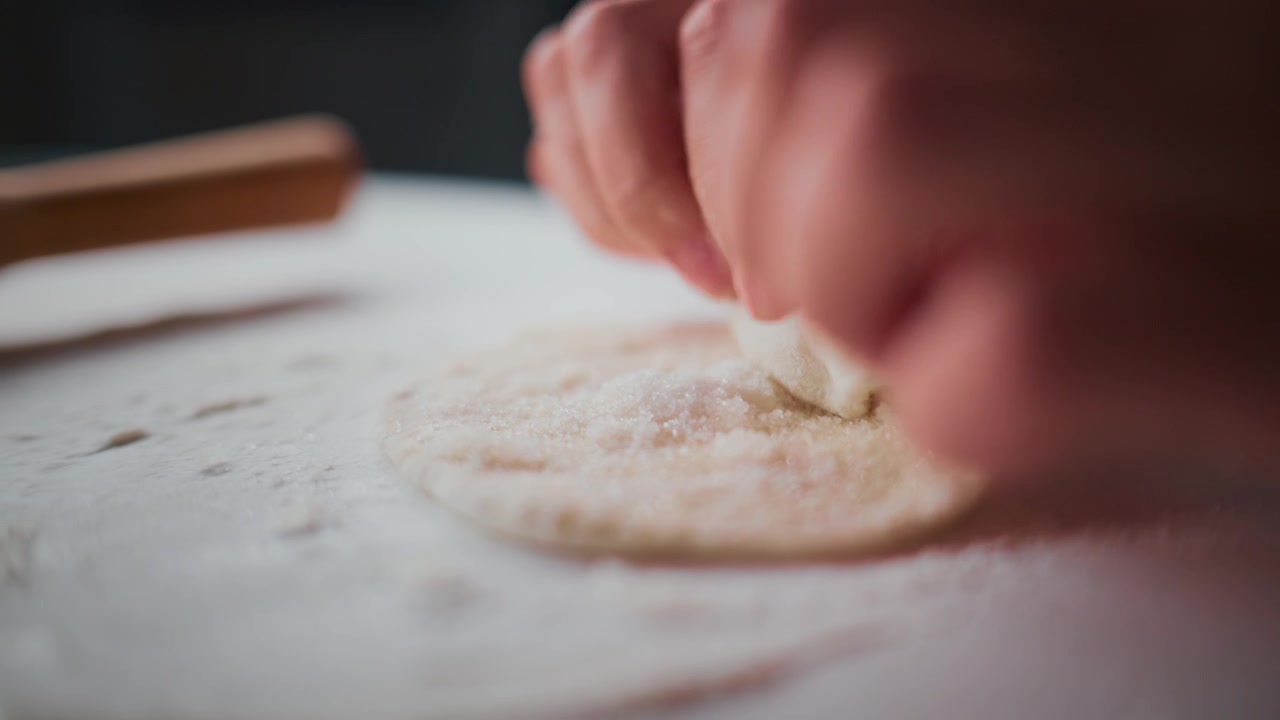 Close-up of hands shaping dough on flour-dusted surface. Fingers press gently, forming round base. Soft lighting highlights texture and motion. Perfect for food tutorials, baking content