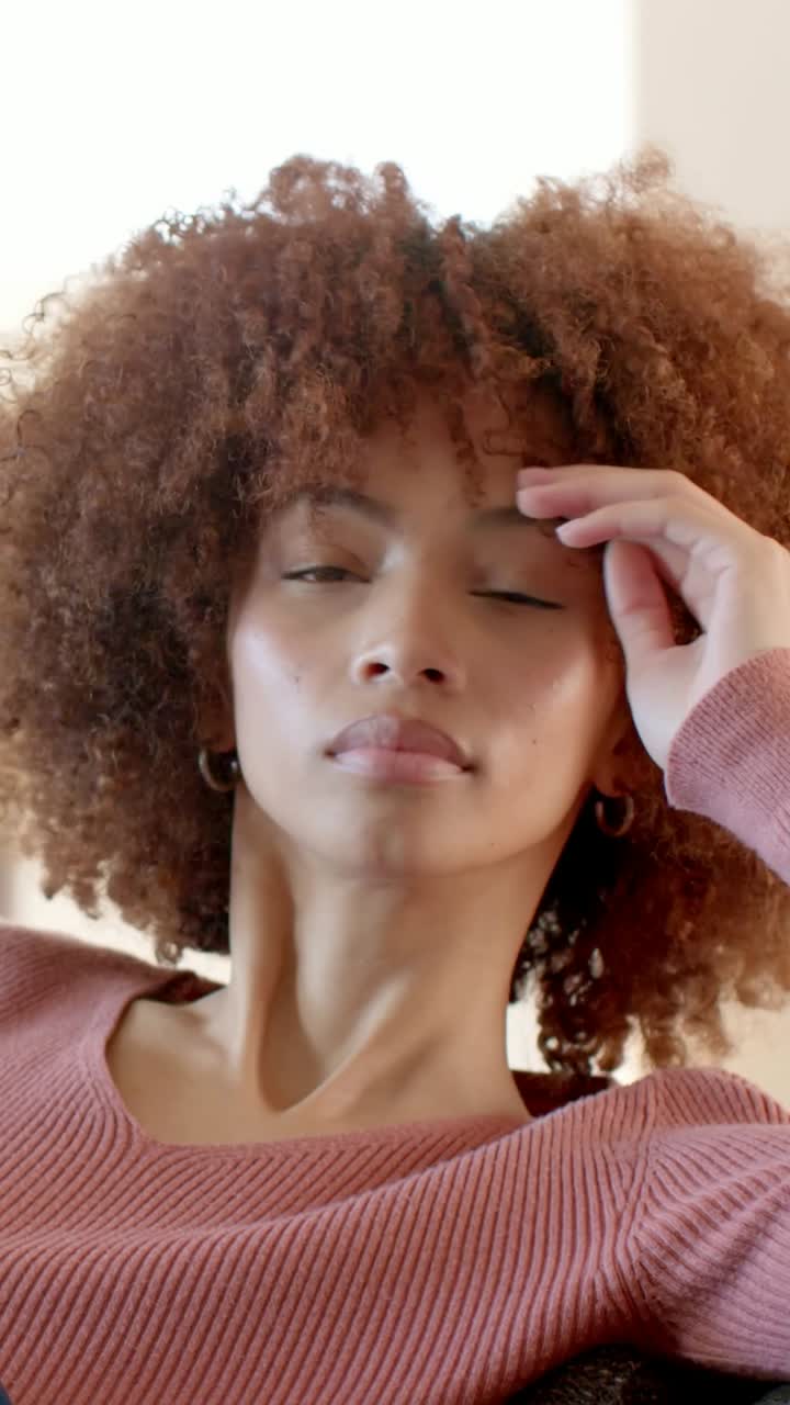 Vertical video: Glancing down at phone, woman in mauve sweater relaxing, adjusting curls on couch