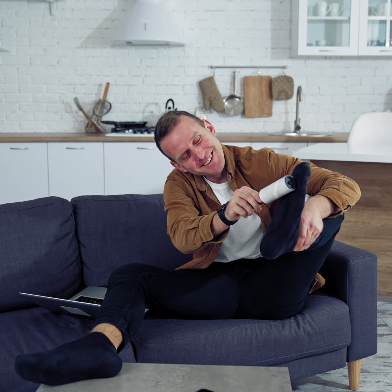 Young man rests on a couch at home. Funny man brushing his socks while sitting on a sofa next to laptop at home. Quarantine concept