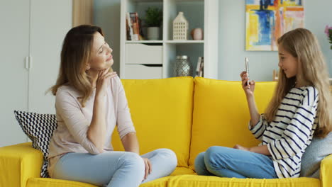 Cute-Blonde-Girl-Taking-Photos-Of-Her-Mother-With-Smartphone-Sitting-On-Yellow-Sofa-In-Living-Room