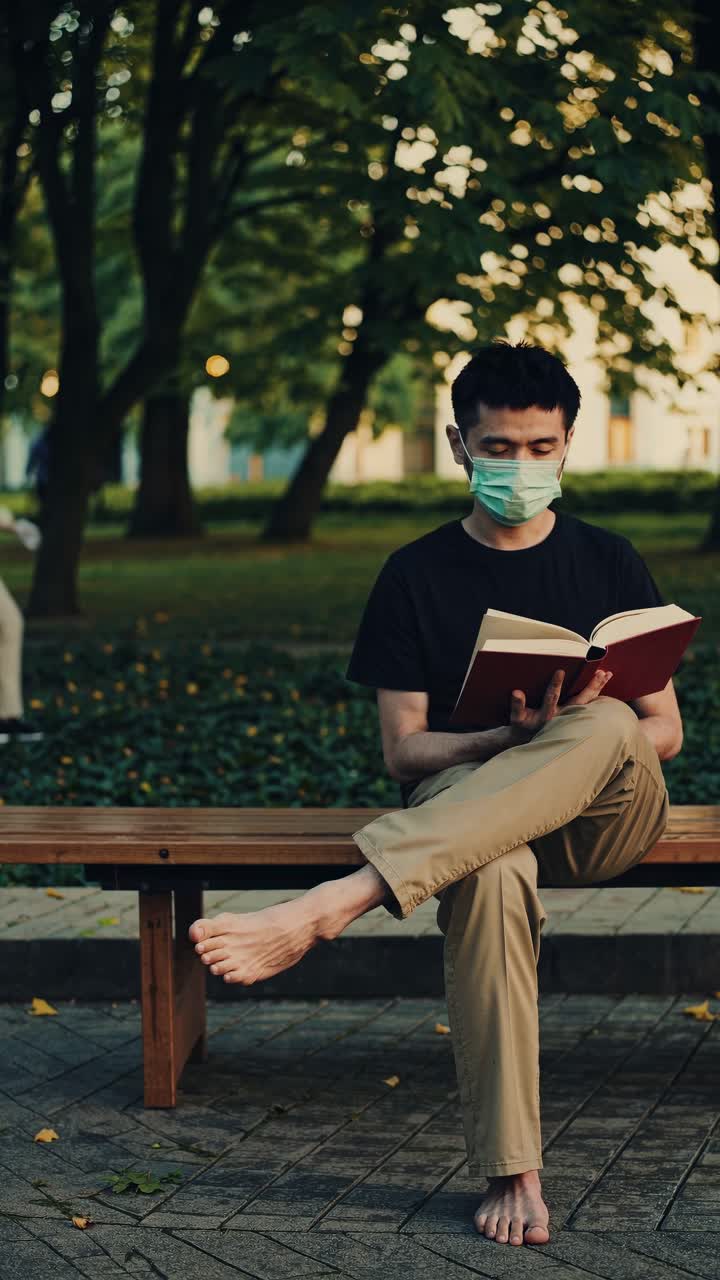 Student sitting barefoot on park bench, reading book while wearing protective face mask, immersed in quiet, peaceful outdoor environment during pandemic period