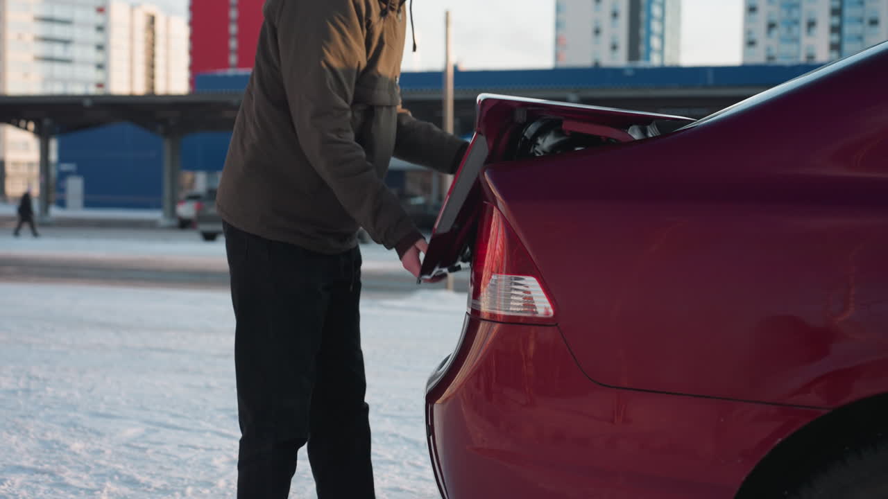Young man in brown jacket walks toward parked red car on snowy ground and reaches to open rear trunk with urban buildings and parked vehicles in background under cold daylight