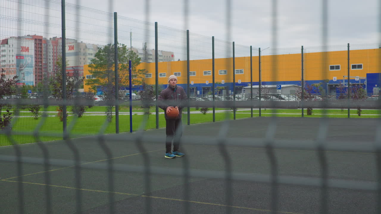 Ung mann som skyter basketball utendørs, overskyet himmel, inngjerdet lekeplass, slowmotion-swish, kontakt med ringen, sprett, treningsdrill, fokusert stilling, joggesko på asfalt, gul industribygning