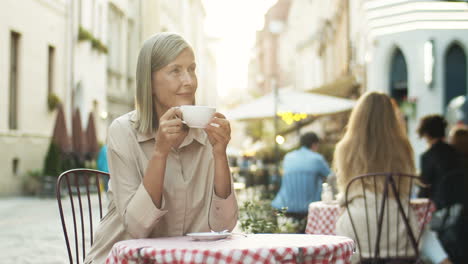 Gut-Aussehende-Seniorin-Lächelte-Frau-Mit-Grauem-Haar,-Die-Kaffee-Schlürfte,-Während-Sie-Draußen-Auf-Der-Caféterrasse-Am-Tisch-Saß