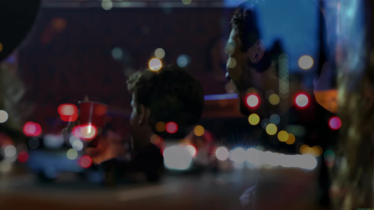 Interieur van een rijdende auto met stadslichten die verschuiven en scherpstellen op twee mannen die een kopje drinken en lachen