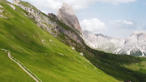 Green-meadow-slope-and-pathway-leading-up-to-Dolomites-of-Italy,-aerial-view