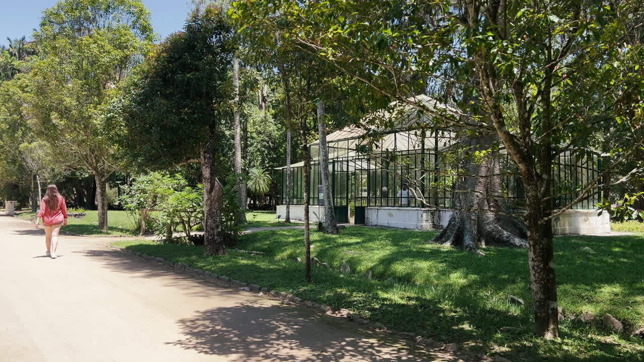 Rear shot of a young woman with long hair, wearing a terracotta-colored shirt, walking on a park path towards the Fern Greenhouse in the Rio de Janeiro Botanical Garden